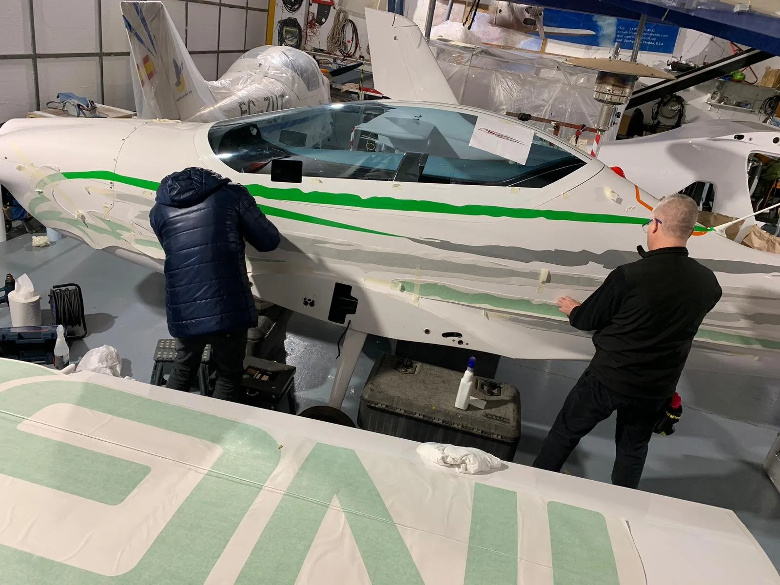 Dos personas aplicando el vinilo verde sobre el fuselaje de un ultraligero en el hangar